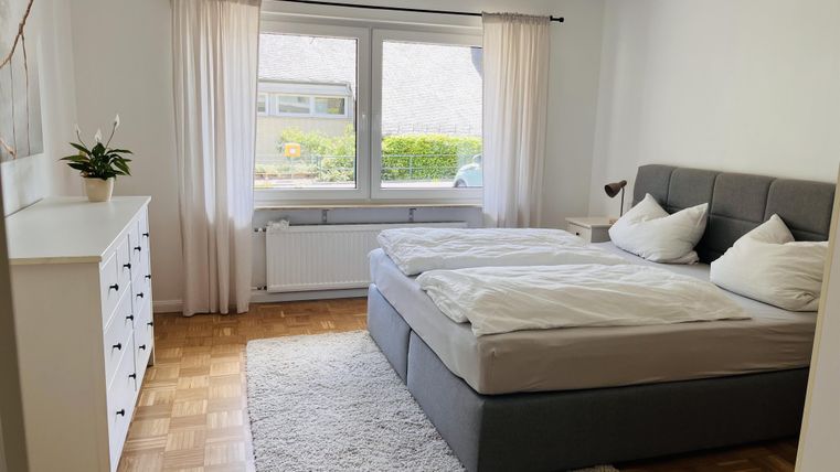 Ein gemütliches Schlafzimmer mit einem großen, bequemen Bett und weißen Bettbezügen. Der Raum hat große Fenster, die viel Tageslicht hereinlassen.