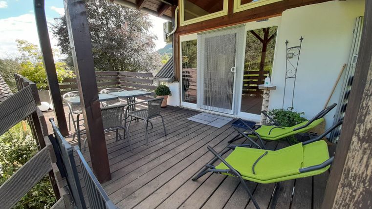 Eine gemütliche Terrasse mit Holzbohlen und modernen Liegestühlen. Im Hintergrund sind ein Tisch und Stühle sowie eine große Glasschiebetür zu sehen.