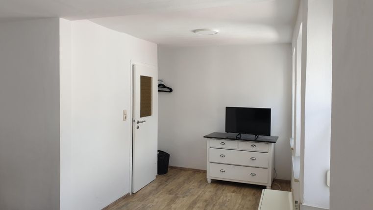 Ein einfaches Zimmer mit einem Fernseher und einer Kommode. Die Wände sind weiß und der Boden ist aus Holz.