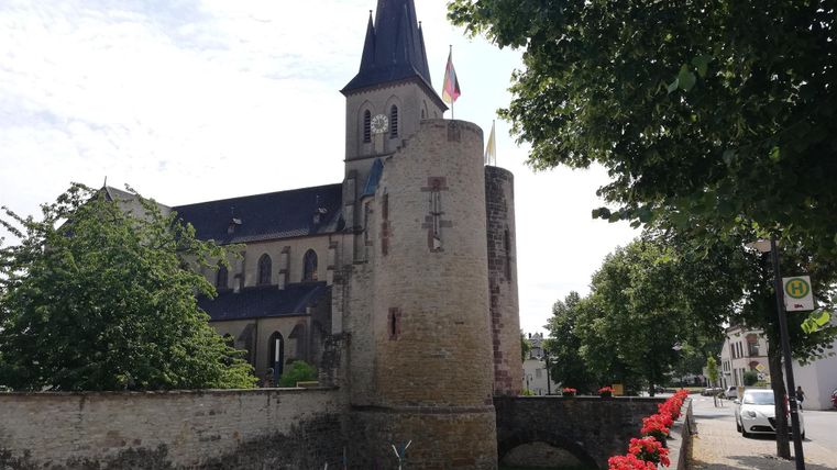 Eine alte Kirche mit einem hohen Turm und einer Mauer aus Stein. Rote Blumen säumen den Weg, während Bäume im Hintergrund Schatten spenden.