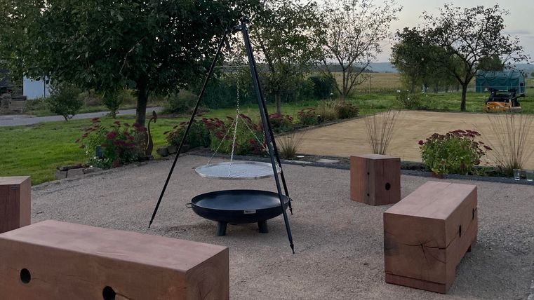 Ein gemütlicher Außenbereich mit einem Lagerfeuerplatz und mehreren Sitzbänken aus Holz. Umgeben von Bäumen und einer grünen Wiese bietet der Ort eine einladende Atmosphäre.