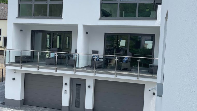 Ein modernes Haus mit großen Fenstern und einem Balkon. Vor dem Haus befindet sich ein gepflasterter Parkplatz mit zwei Garagen.