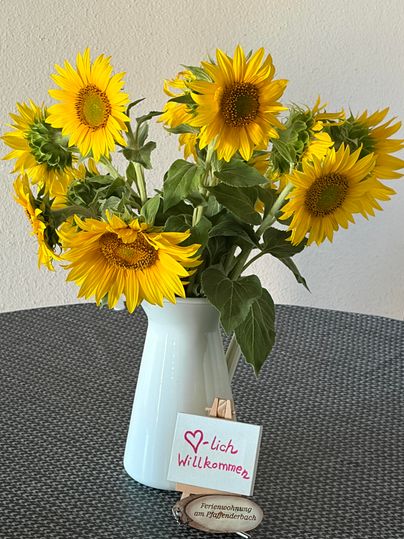 Ein Strauß Sonnenblumen in einer weißen Vase steht auf einem Tisch. Darunter befindet sich ein Willkommensschild.