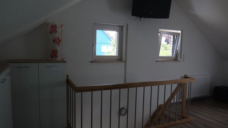 Ein gemütlicher Raum im Dachgeschoss mit einem kleinen Geländer und Fenstern. An der Wand hängt ein Fernseher, und es gibt einen Schrank sowie einen Blick auf die Umgebung.