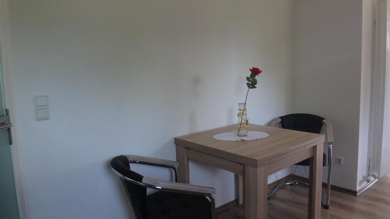 Ein heller Raum mit einem Tisch und zwei Stühlen. Auf dem Tisch steht eine Rose in einer Vase.