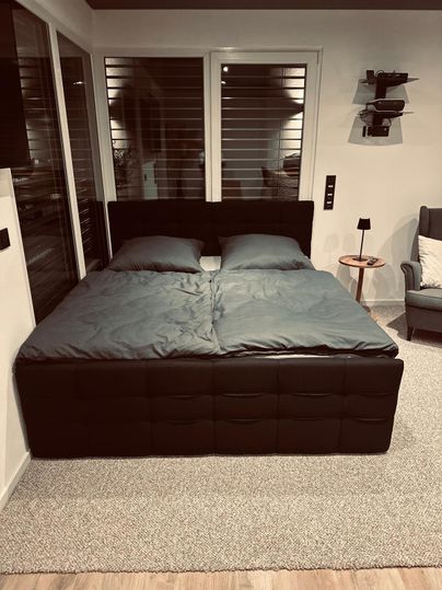 Ein modernes Schlafzimmer mit einem großen Doppelbett und grauer Bettwäsche. Neben dem Bett steht ein kleiner Tisch und ein Sessel.