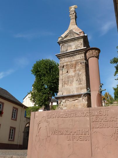 Ein historischer Obelisk steht in einer kleinen Stadt. Umgeben von Bäumen und Häusern, zeigt er lokale Architektur und Geschichte.