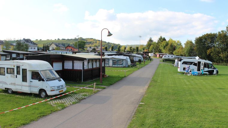 Ein Campingplatz mit verschiedenen Wohnmobilen und Zelten. Die Landschaft ist grün und es gibt einen gepflasterten Weg zwischen den Stellplätzen.