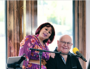 Ein fröhliches Paar steht vor einem Fenster und lächelt in die Kamera. Im Hintergrund sind Mikrofone und eine schöne Aussicht zu sehen.
