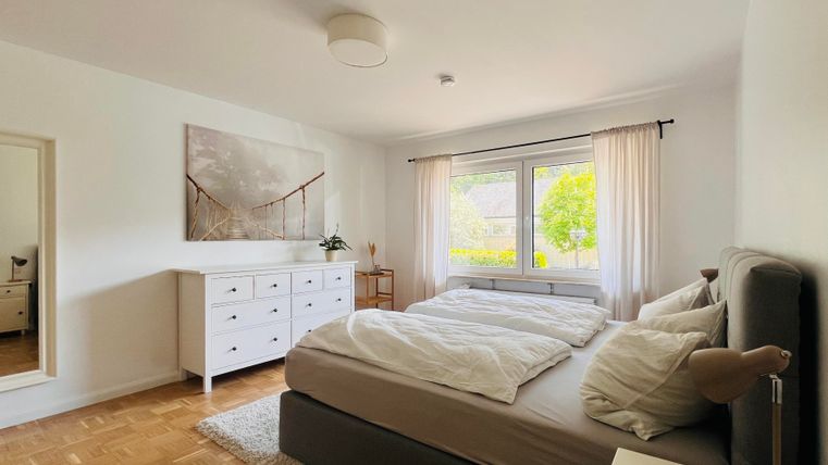 Ein modernes Schlafzimmer mit einem großen Bett und hellen Vorhängen. Es gibt eine Kommode und ein Fenster, das auf einen grünen Außenbereich blickt.