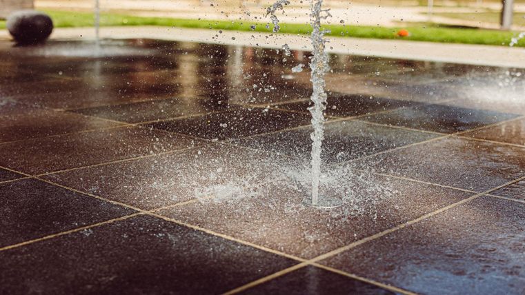 Ein Wasserstrahl spritzt aus dem Boden auf einem glatten Platz. Die Umgebung wirkt hell und einladend.