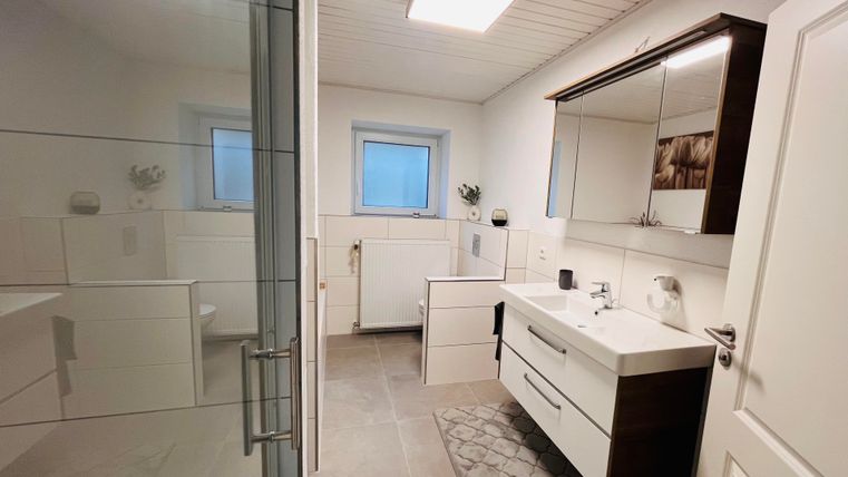 Ein modernes Badezimmer mit einer Dusche aus Glas und weißen Möbeln. Der Raum ist hell und freundlich, mit einem großen Spiegel über dem Waschbecken.