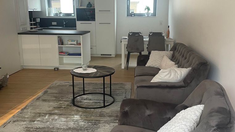 Die großzügige Fensterfront macht das Wohnzimmer besonders lichtdurchflutet und freundlich. Das gemütlich Sofa, ein bequemer Sessel sowie der Fernseher auf dem Sideboard laden zum Entspannen ein. Kissen und Decken sorgen für zusätzlichen Komfort.