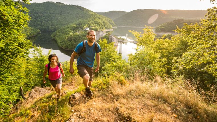 Zwei Wanderer auf einem Pfad mit Blick auf einen Stausee und bewaldete Hügel im Hintergrund.
