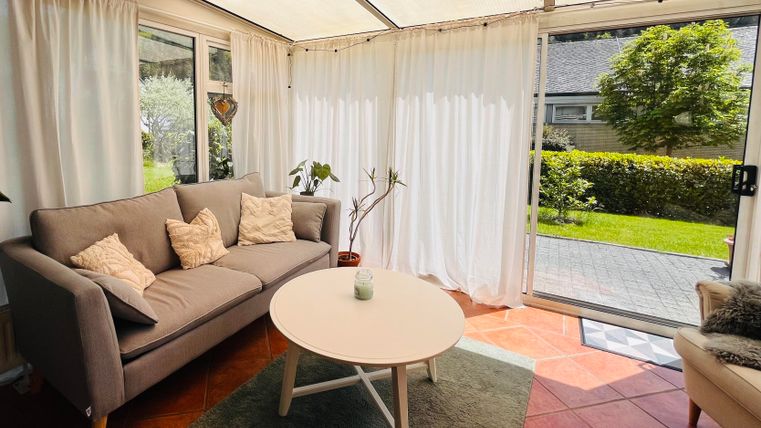 Ein helles Wohnzimmer mit einem grauen Sofa und einem runden Tisch. Große Fenster lassen viel Tageslicht herein und bieten Blick ins Grüne.