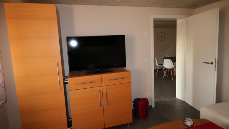 Ein modernes Wohnzimmer mit einem Fernseher auf einem Holzschrank. Im Hintergrund ist ein Essbereich mit weißen Stühlen zu sehen.