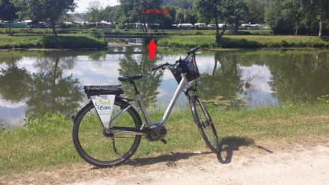 Ein Fahrrad steht am Ufer eines ruhigen Gewässers. Im Hintergrund sind Bäume und ein Campingplatz zu sehen.