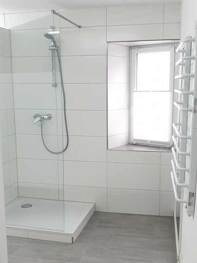 Ein modernes Badezimmer mit einer Glasdusche und hellen Fliesen. Der Raum hat ein Fenster und eine Handtuchheizung.