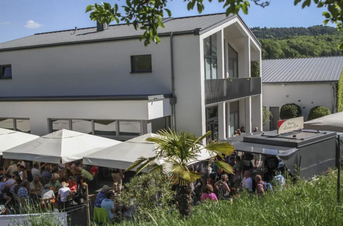 Eine moderne Veranstaltungslocation mit vielen Gästen im Freien unter Sonnenschirmen. Der Ort ist umgeben von Grün und hat eine einladende Atmosphäre.
