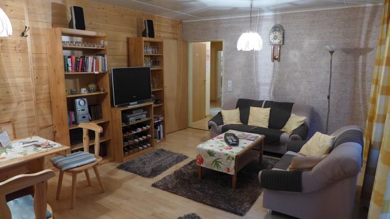 Ein gemütliches Wohnzimmer mit einem Fernseher, einem Sofaset und einem Esstisch. Helle Wände und Holzmöbel sorgen für eine warme Atmosphäre.