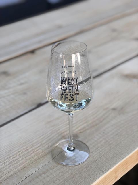 Ein Glas mit dem Aufdruck "WEST WEINFEST" steht auf einem Holztisch. Der Tisch hat eine natürliche Holzoberfläche und schafft eine gemütliche Atmosphäre.