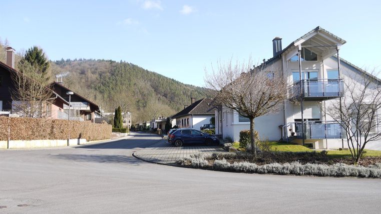 Eine ruhige Wohnstraße mit modernen Häusern und Bäumen. Im Hintergrund sind sanfte Hügel unter einem klaren Himmel zu sehen.