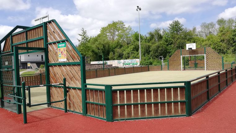 Ein Sportplatz mit einem eingezäunten Bereich für Basketball und andere Spiele. Die Umgebung ist grün und einladend.