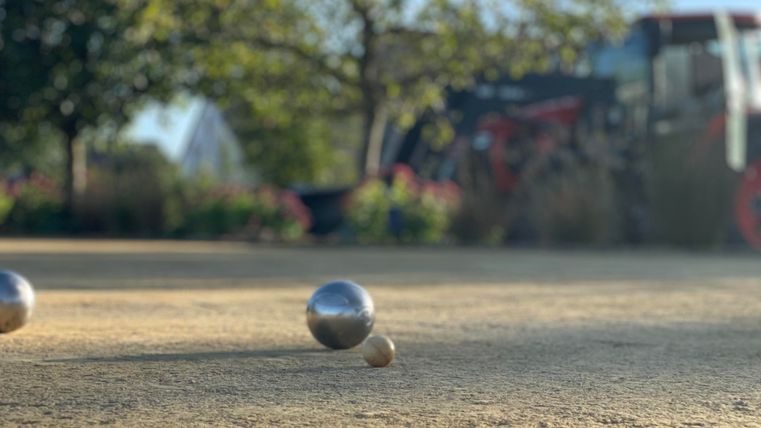 Drei Metallkugeln liegen auf einem Sandplatz. Im Hintergrund sind grüne Bäume und ein roter Traktor zu sehen.