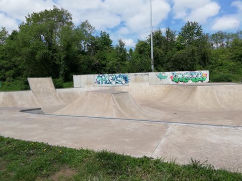 Ein Skatepark mit verschiedenen Rampen aus Beton. Im Hintergrund sind Bäume und Graffiti-Wände zu sehen.