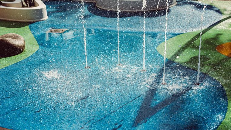 Ein bunter Wasserspielplatz mit sprudelnden Wasserfontänen. Der Boden ist in verschiedenen Farben gestaltet und vermittelt eine fröhliche Atmosphäre.