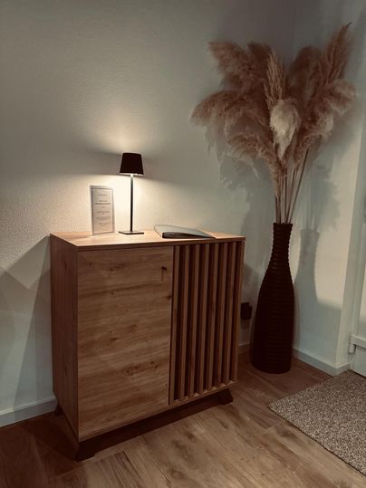 Ein stilvoller Holzschrank steht neben einer dekorativen Lampe. Im Hintergrund befindet sich eine große Vase mit Trockenblumen.