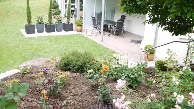 Ein gepflegter Garten mit bunten Blumen und grünen Pflanzen. Im Hintergrund steht ein moderner Sitzbereich mit Stühlen und Tischen.