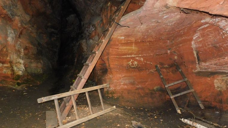 Eine Holzleiter steht neben einer roten Felswand in einer Höhle. Der Boden ist mit Laub und kleinen Ästen bedeckt.