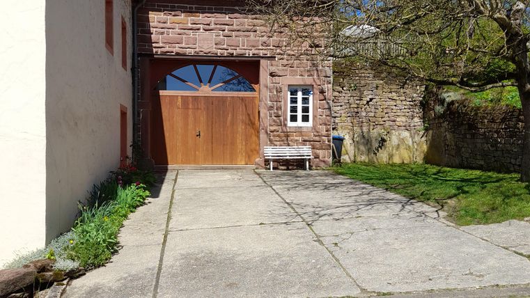 Ein charmantes Gebäude mit einer Holztür und einem gepflasterten Vorplatz. Die Umgebung ist grün und es ist ein klarer blauer Himmel zu sehen.