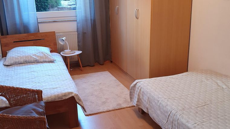 Ein einfaches Schlafzimmer mit zwei Betten und einem großen Kleiderschrank. Vor dem Fenster sind Vorhänge und ein kleiner Tisch steht neben einem der Betten.
