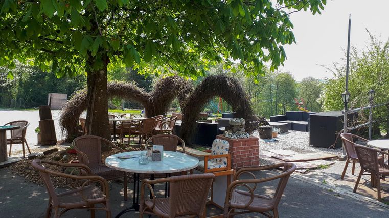 Ein gemütlicher Außenbereich mit Tischen und Stühlen unter einem Baum. Im Hintergrund sind dekorative Elemente und eine sonnige Atmosphäre zu sehen.