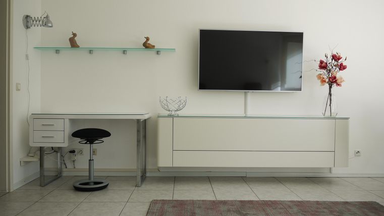 Ein modernes Wohnzimmer mit einem weißen Fernseher, einem Schreibtisch und einer stilvollen Blumendekoration. Die Wände sind hell und der Boden ist mit Fliesen ausgelegt.