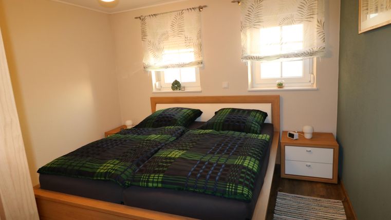 Ein gemütliches Schlafzimmer mit einem Doppelbett und grünen Bettdecken. Zwei Fenster lassen viel Licht herein und ein Nachttisch rundet die Einrichtung ab.