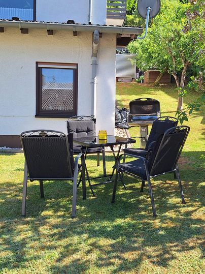 Ein schöner Tisch mit Stühlen auf dem Rasen neben einem Haus. Im Hintergrund ist ein Grill und ein Baum zu sehen.