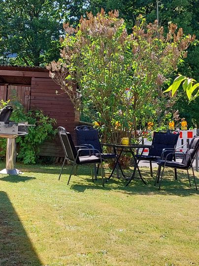 Ein einladender Garten mit einem Tisch und Stühlen unter einem Baum. Im Hintergrund steht ein Holzhaus und die Sonne scheint.
