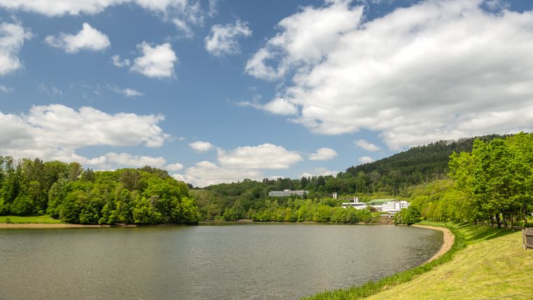 Stausee Bitburg mit bewaldeten Hügeln und Gebäuden im Hintergrund unter blauem Himmel mit Wolken.