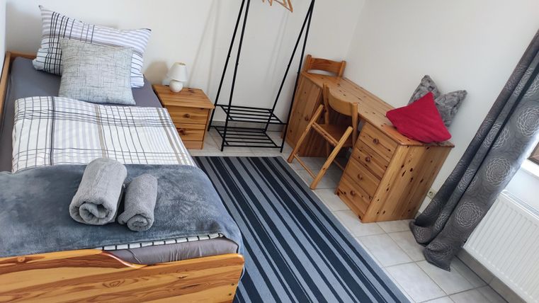 Ein einfaches und gemütliches Zimmer mit einem Einzelbett, einem Schreibtisch und einer Garderobe. Der Raum hat helle Wände und einen modernen Teppich.