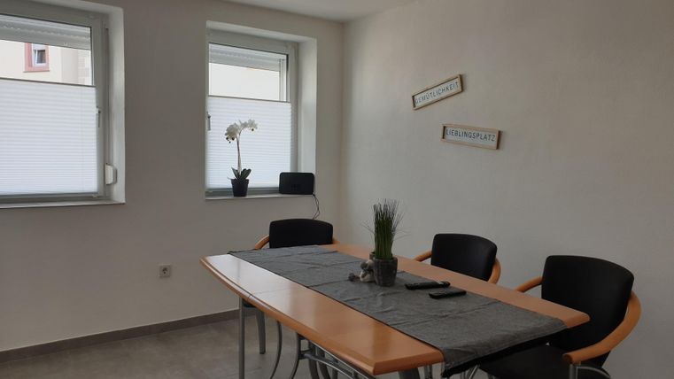 Ein heller Raum mit einem Tisch und schwarzen Stühlen. Auf dem Tisch steht eine Pflanzen dekoriert mit einer Tischdecke.