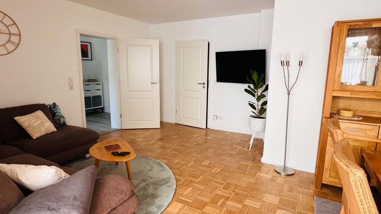 Ein gemütliches Wohnzimmer mit einem grauen Sofa und einem runden Tisch. Es gibt einen Holzschrank, einen Fernseher an der Wand und viel Licht durch die Fenster.