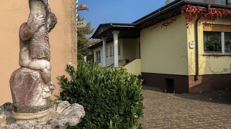 Eine dekorative Statue steht vor einem Gebäude mit gelber Fassade und grünen Büschen. Der Weg führt zu einem freundlich aussehenden Hauseingang.