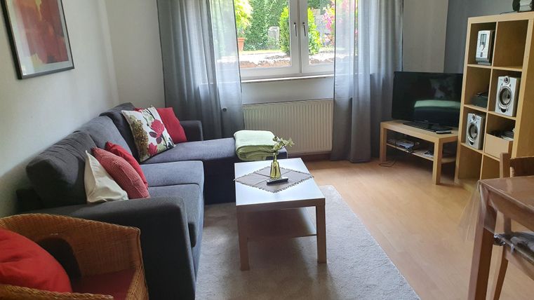 Ein gemütliches Wohnzimmer mit einem grauen Sofa, einem Holz-Esstisch und einem Fernseher. Große Fenster lassen viel Tageslicht herein und verleihen dem Raum eine einladende Atmosphäre.