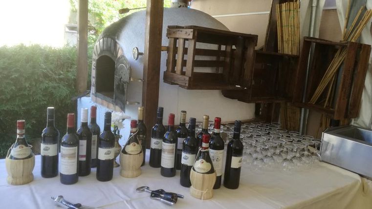 Ein schön gedeckter Tisch mit verschiedenen Rotweinen und Gläsern. Im Hintergrund befindet sich ein traditioneller Pizzaofen.