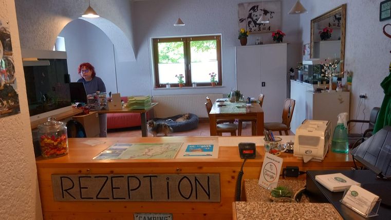 Ein Empfangsbereich mit einem Holztresen und der Aufschrift „REZEPTION“. Hinter dem Tresen sitzt eine Person und es gibt Tische und Stühle im Raum.