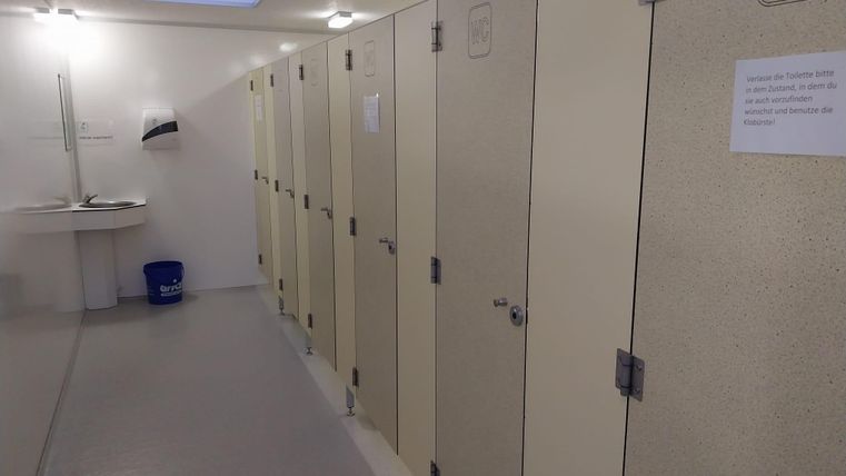 Ein sauberer Toilettenbereich mit mehreren Kabinen. An der Wand hängt ein Handwaschbecken und ein Hinweisblatt.