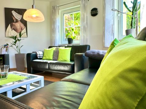 Ein gemütliches Wohnzimmer mit einem schwarzen Sofa und bunten Kissen. Das Licht fällt durch ein Fenster und es gibt eine Pflanze und ein Kunstwerk an der Wand.